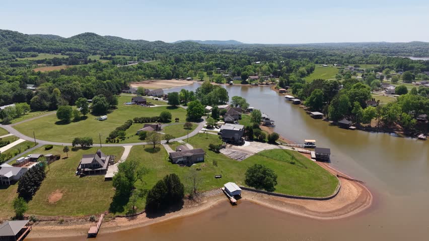 Low altitude drone flyover of the Georgetown Tennessee rural community featuring homes green lawns vehicles and a dirt road with the Hiwassee River curving through the right and distant wooded ridges