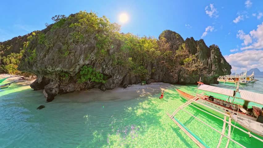 Entalula Beach in El Nido, Philippines, limestone cliffs, vibrant tropical vegetation, and traditional outrigger boats floating on crystal-clear turquoise waters under sunny blue sky of Palawan island