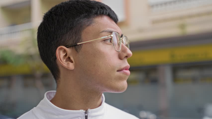 Man with round gold glasses and visible earring looking at camera with neutral expression, white zip jacket on street; contemplation identity confidence.