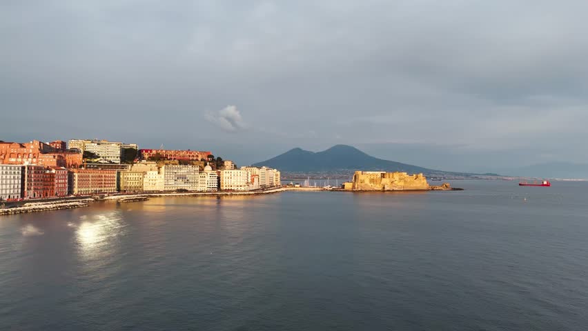 Aerial view of Naples, Italy, with Castel dell’Ovo, Mount Vesuvius, and calm bay water lit by soft evening reflections along the historic seafront.