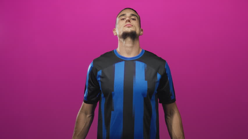 Man in blue and black soccer jersey holds index fingers to head like horns in studio, hands visible and direct stare; playful taunt.