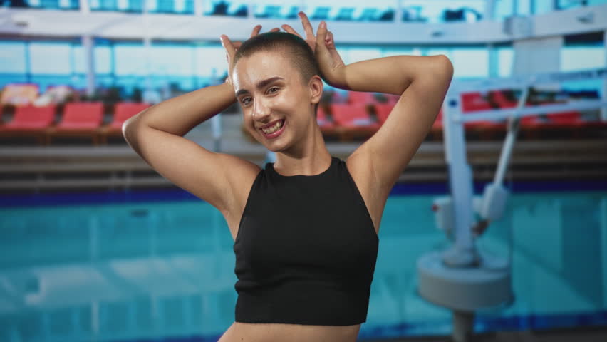 Woman with buzzcut and bare midriff posing with hands to head making playful horns, smiling and dancing in a building; joy playfulness.