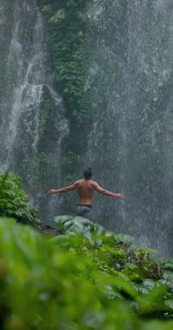 Handsome man with raised arms under powerful waterfall, feeling energy in tropical jungle nature
