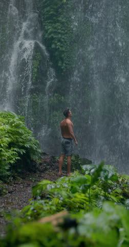 Handsome man under powerful waterfall, feeling energy in tropical jungle nature