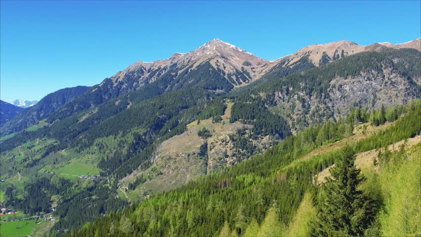 Aerial view of Bad Gastein, Austria, showing an alpine valley with villages, forests, green meadows, winding roads, and surrounding mountain peaks under a clear blue sky.