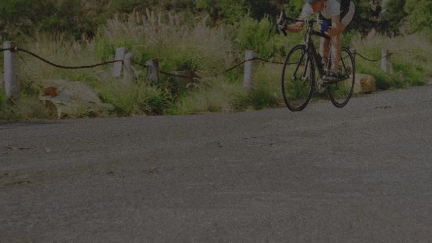 Woman cyclist entering frame, pedaling on rope-fence road, low angle showing wheel spin for fitness. Rider, bike, outdoors, sunny, motion, shadows, rural