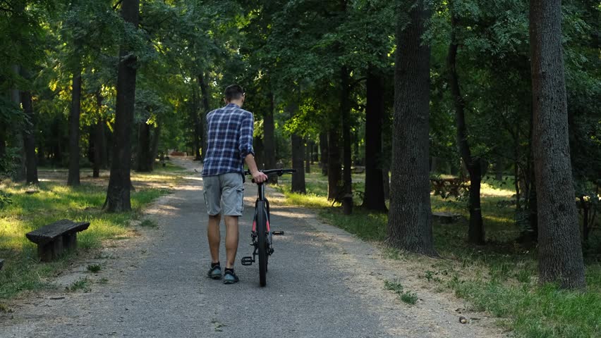 A man walks through a forest park pushing a bicycle beside him. Sun rays shine through the trees creating a peaceful atmosphere
