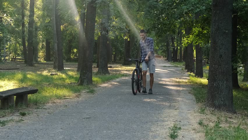 A man walks through a forest park pushing a bicycle beside him. Sun rays shine through the trees creating a peaceful atmosphere