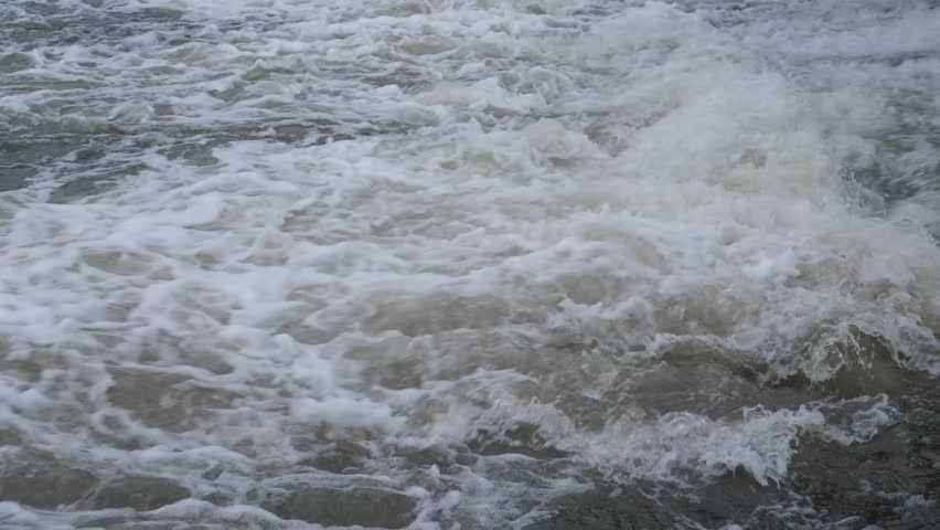 Rushing water stream with white foam and strong current. Close-up of powerful river rapids or spillway on an overcast day