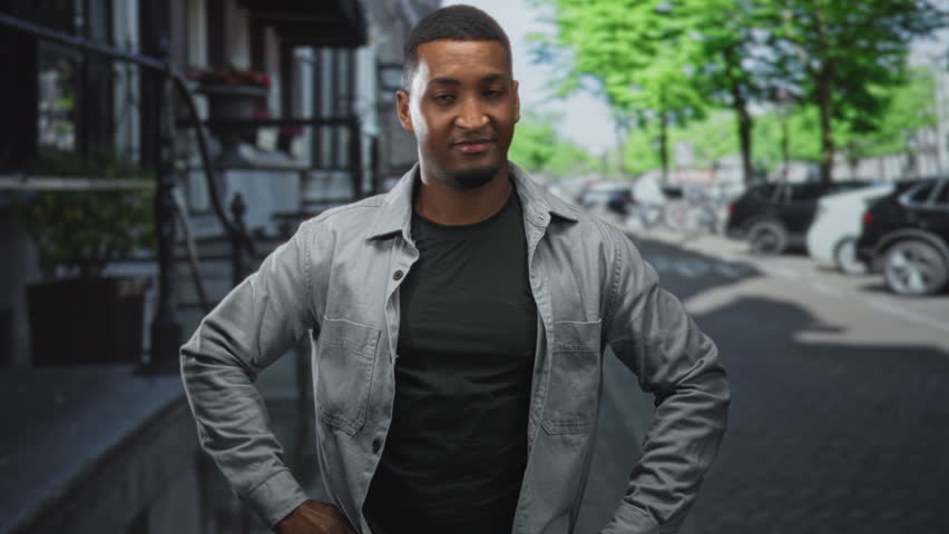 Young black man posing with hands on hips, wearing jacket and black tshirt on street; confidence approachability warmth.