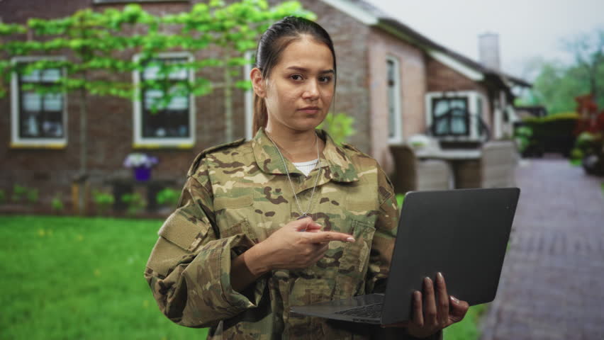 Woman soldier in camouflage holds a laptop with dog tags visible while standing on a brick path by a building; duty resilience.