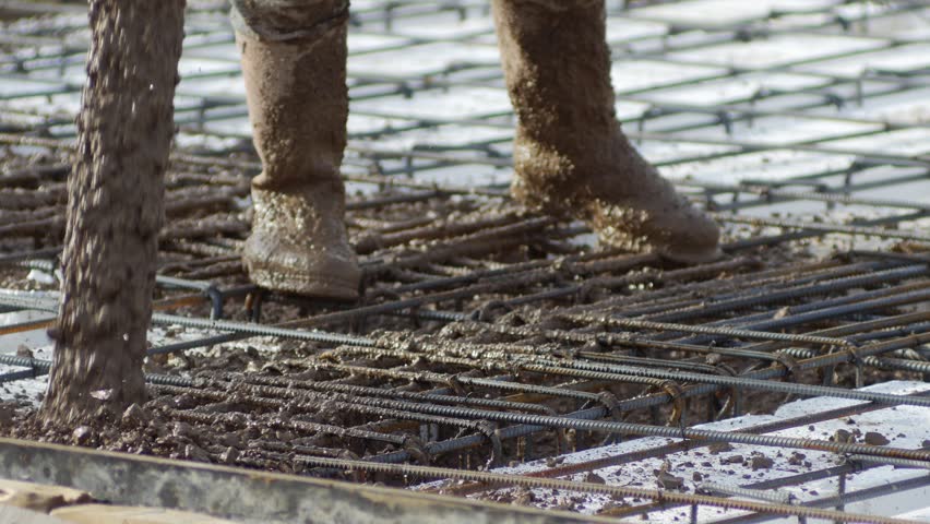 Close-up of concrete-covered rubber boots standing on rebar grid during concrete pouring, wet concrete splash and reinforcement mesh detail at construction site