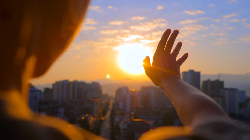 Close up: silhouette of woman is waving her hand against the sunset, sunrise sky over the city, sun is shining through fingers: over the shoulder view, sun lens flares. Freedom and inspiration concept