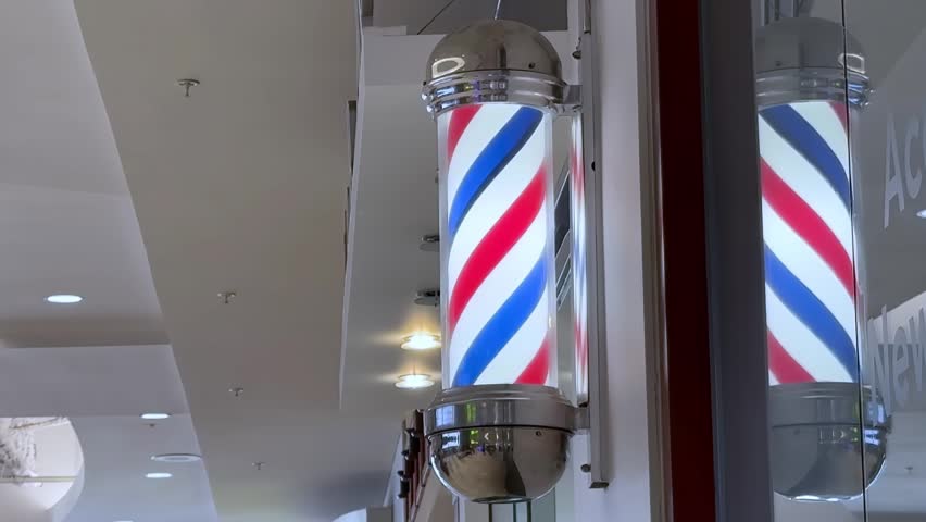 Red, white, and blue illuminated barbershop pole mounted on a wall in a modern indoor shopping center with a clean, bright aesthetic.