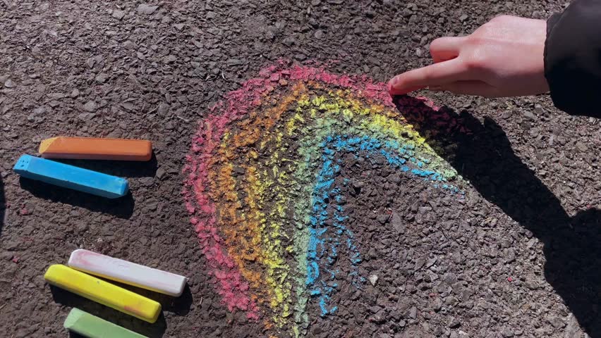Drawing of a rainbow with chalk on the asphalt with colorful pieces of chalk lying nearby. Children