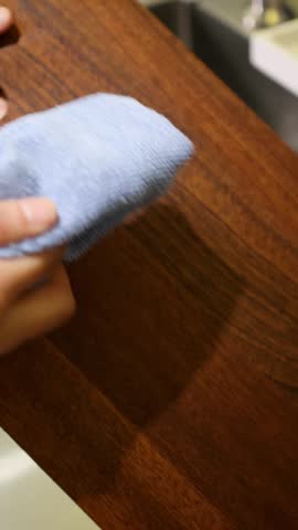 Close-up shot of hands drying a wet wooden cutting board using a blue microfiber cloth