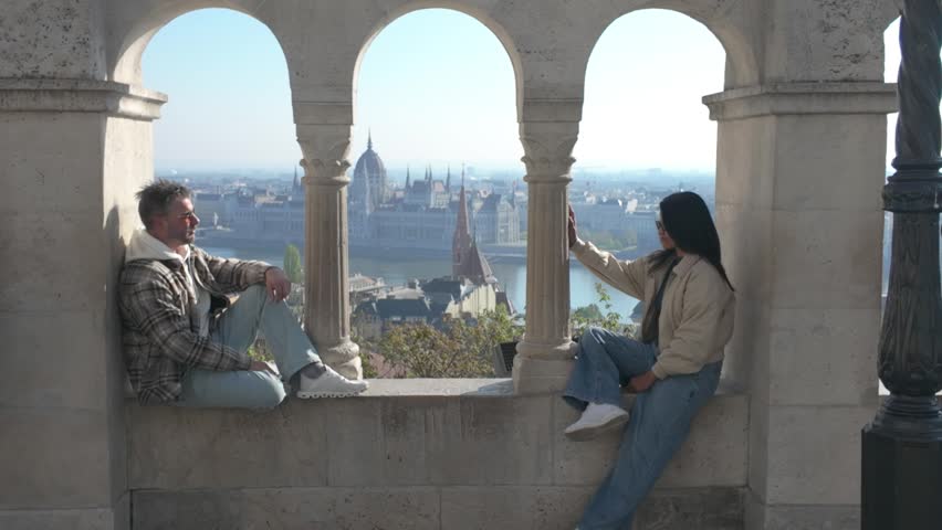 A couple relaxes at Fishermans Bastion, gazing at stunning panoramic views of Budapest. Sunlight illuminates the historic architecture as they share joyful moments together, surrounded by history.