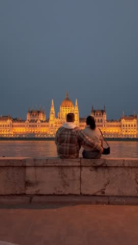 Couple relaxes together on a stone wall in Budapest, enjoying the stunning view of the citys historic buildings lighted up at dusk, creating a romantic and tranquil atmosphere.