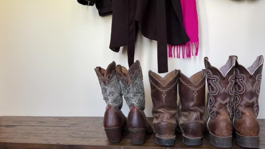 Three pairs of worn leather cowboy boots arranged in size order on a wooden shelf evoke the warmth of Australian rural family life, with a dark jacket and pink scarf hanging above suggesting a mudroom entryway in a country home.