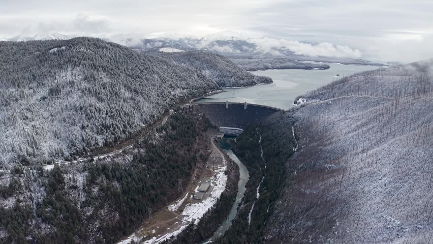 Aerial drone view of snow-dusted hills with a winding road and river near Kicking Horse Dam, Montana.