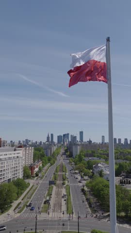 Drone aerial shots of a large Polish flag waving dynamically in the wind, captured from medium distance with Warsaw city center buildings as backdrop. Flag mounted on tall flagpole atop public building or rooftop—white over red fabric billows and snaps crisply, edges frayed slightly, pole guy-wires taut. Sunlight filters through fabric folds, casting shadows on taut sections