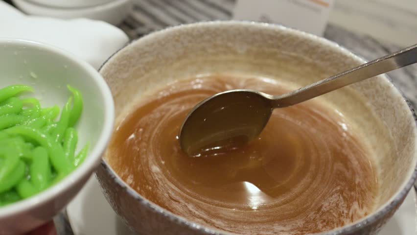 Close-up shot of pouring brown coconut milk syrup over green rice flour noodles in bowl
