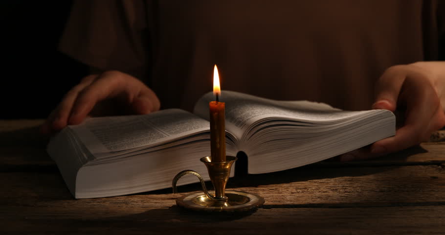 Woman reading Bible at wooden table with burning candle, closeup