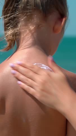 Mothers hands apply protective sunscreen on daughters skin back on background of beach with blue sea. Skin care from sun. Summer vacation travel holiday. Vertical shot
