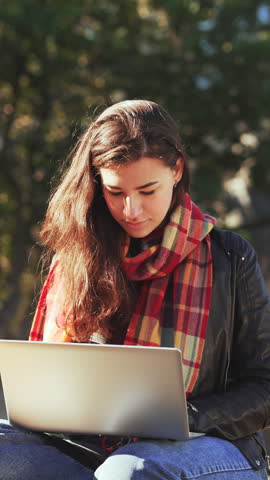 Happy brunette girl in scarf sitting with laptop and looking away with smile in fall park. Inspired female freelancer working outdoors