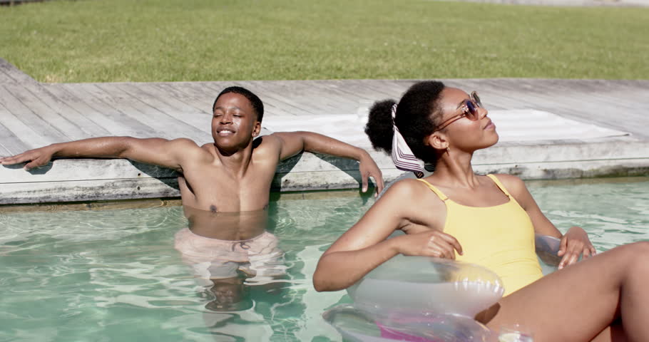 African American couple relaxing at pool sun making woman in yellow suit adjusting shades on float. Waterside, pair, lifestyle, sunshine, backyard, inflatable, sunglasses