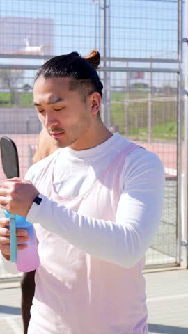 Young asian man with a man bun drinking from a reusable water bottle to rehydrate after an intense pickleball match, feeling refreshed and smiling on a sunny day at an outdoor court