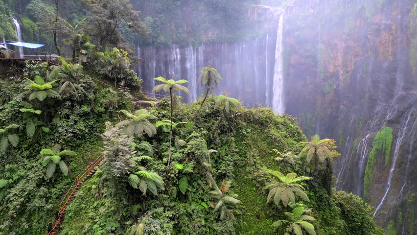 Aerial drone view of Tumpak Sewu waterfall in East Java, Indonesia. Spectacular curtain waterfall surrounded by dense tropical jungle and cliff edge with visible trail.