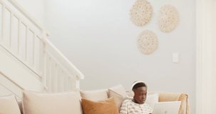 African youth male settling into couch, typing on laptop, wearing headphones and maintaining focus. Computer, sofa, livingroom, striped, shirt, shorts, plant - Powered by Shutterstock - Get 15% off with code: PIKWIZARD15