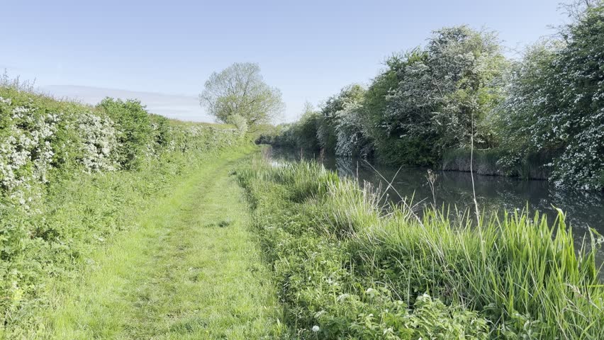 Looking along a quaint and picturesque canal in rural England. There is greenery on both sides of the canal, and the water is still against a blue sky.