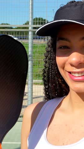 Beautiful mixed race woman with curly hair smiling and holding a paddle on a sunny pickleball court. The female athlete enjoys a summer day playing her favorite sport outdoors