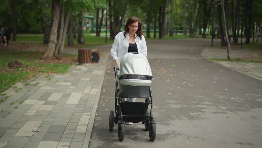 White mother pushing stroller through park, gentle paced walk on paved path, leafy trees lining walkway, soft overcast light, casual jacket, baby cocooned under canopy, peaceful parental routine