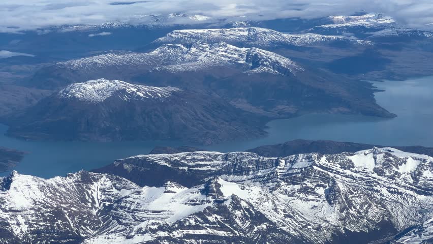 Panoramic aerial view showing majestic mountain peak covered with bright winter snow beside deep blue lake water conveying serene awe inspiring outdoor nature landscape flight under cloudy sky