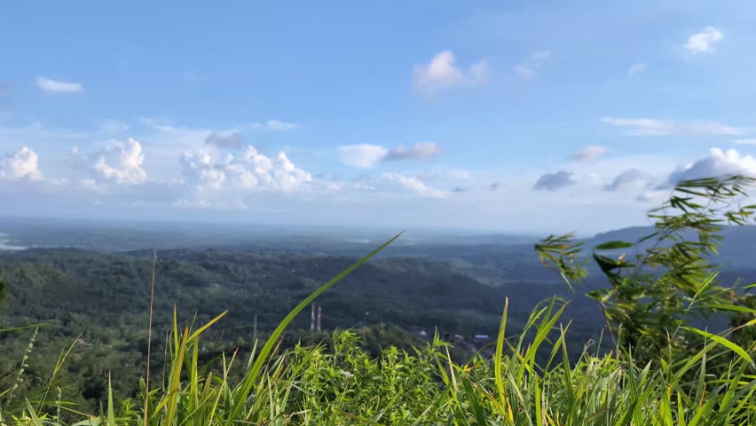 A scenic hilltop view overlooking a rural village and surrounding natural landscape from above. Small houses and fields appear distant below, creating a sense of scale and depth. In the foreground, grass sways gently in the wind, adding movement to the peaceful countryside panorama under daylight.
