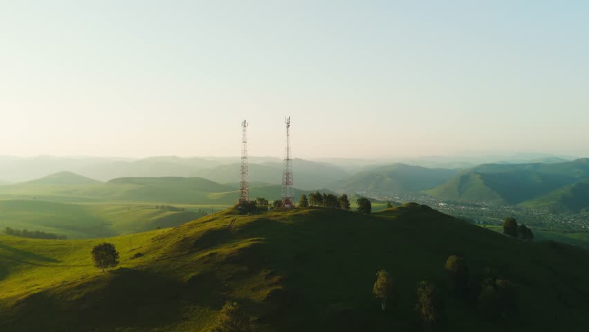 Aerial view of two radio towers atop a lush green hill. Rolling hills stretch into distance under soft morning light. Scenic landscape ideal for nature, tech, or travel projects