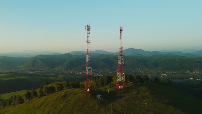 Aerial view of two tall red and white communication towers on a green hill. Rolling hills and valleys extend into the distance. Soft morning or evening light bathes the scene. No people visible