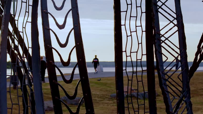 Serene lakeside scene viewed through intricate metal framework. People walk along the waterfront path, evoking sense of contemplation and solitude. Subdued lighting and calm waters create introspectiv