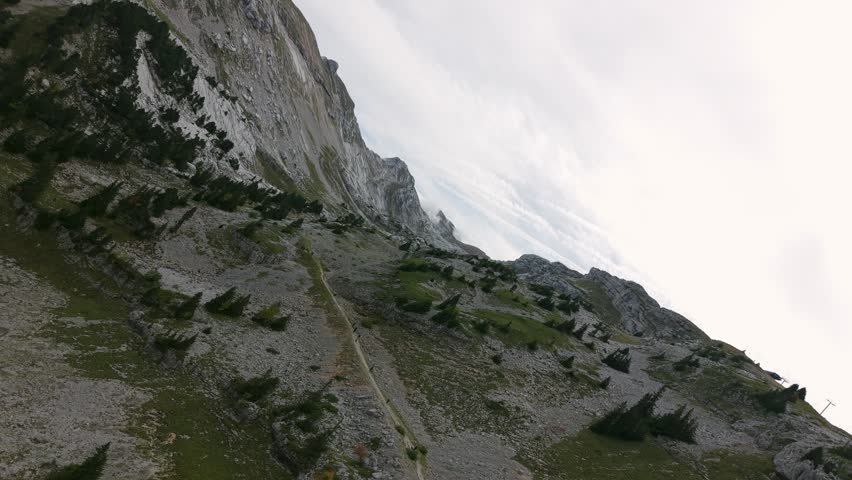 Aerial rotating shot of a rocky alpine ridge with a winding hiking trail, scattered conifers and cloudy sky.