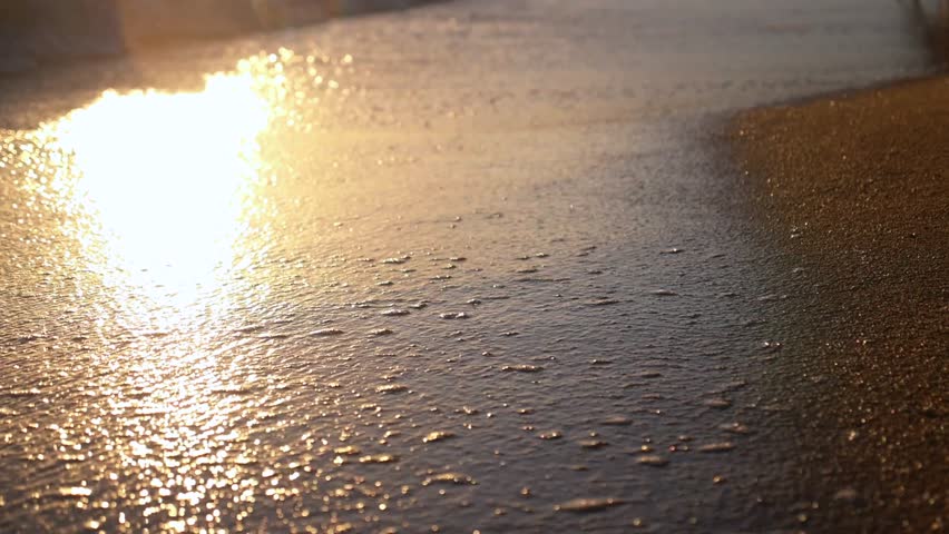 Calm sea water with small waves washing over a sandy beach during a beautiful golden sunset, creating a relaxing and peaceful atmosphere with sparkling sun reflections on the wet sand