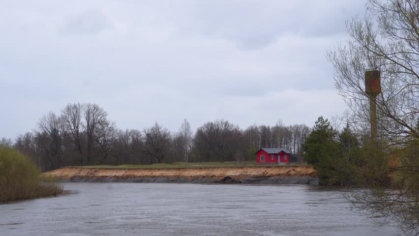 A spring landscape with a river and a red house. A small house stands on the shore, with water and vegetation all around.