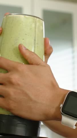 Vertical video: Placing man locking, running blender cup in kitchen, orange tee making smoothie. Forearms, grip, glass, countertop, green, natural, vertical video