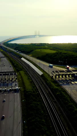 Vertical video. Malmo, Sweden. The Oresund Bridge toll plaza sits at the Swedish approach to the combined road and rail link connecting Sweden and Denmark. The cable-stayed span stretches across the Oresund strait toward the horizon, where its tall pylons and suspended deck catch the warm light of late afternoon. The toll station handles a steady flow of cars and trucks, while the parallel railway runs along the central embankment. Lush green hills and coastal vegetation frame the infrastructure, with the calm sea reflecting sunlight in the background. The structure transitions from span to tunnel via the artificial island Peberholm, an engineering solution chosen to preserve airspace near Copenhagen Airport and maintain open shipping channels. Opened in 2000, the crossing carries both motorway and railway traffic on separate levels, making it the longest combined road and rail span in Europe. The scene captures a functional transportation hub integrated into a serene coastal landscape under clear summer skies. Aerial View, Departure of the camera. Rich colors