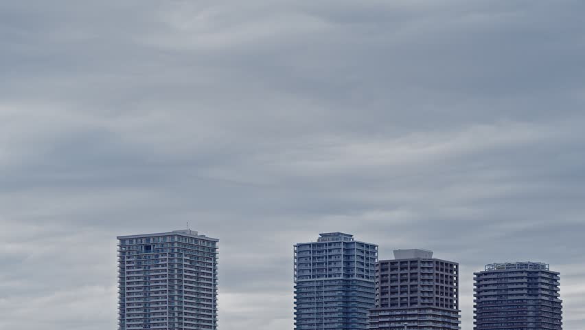 A static wide-angle time-lapse showing thick, textured grey clouds passing over a row of modern high-rise apartment buildings in an urban area.