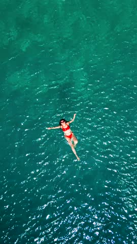 Woman in a red swimsuit swims on her back in the clear sea, top down view