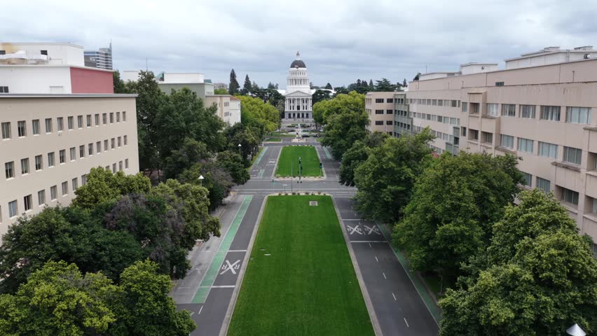 Aerial establishing shot of Sacramento, California, showing State Capitol building, tree lined boulevard, government offices and dramatic cloudy skies in downtown USA. Panorama approaching shot.