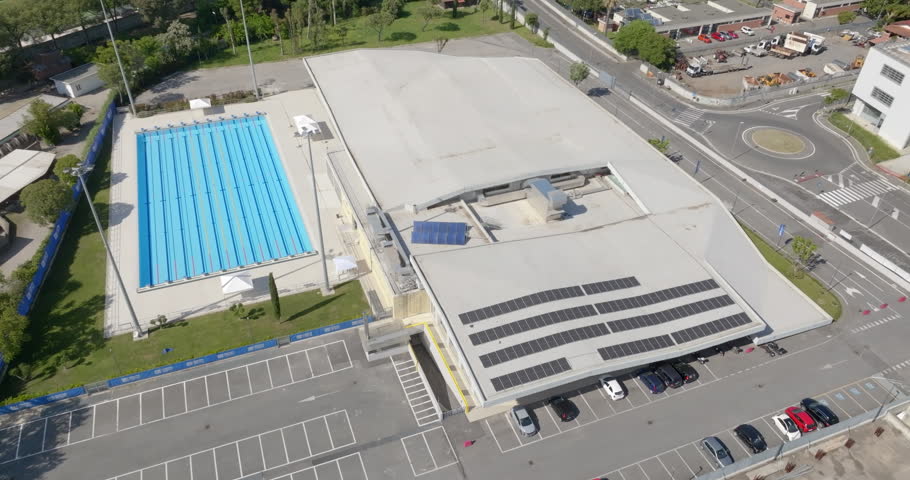 Aerial view of a professional swimming facility featuring a large blue lap pool with marked lanes, situated next to a modern sports building and a spacious parking area. The sport center is empty.